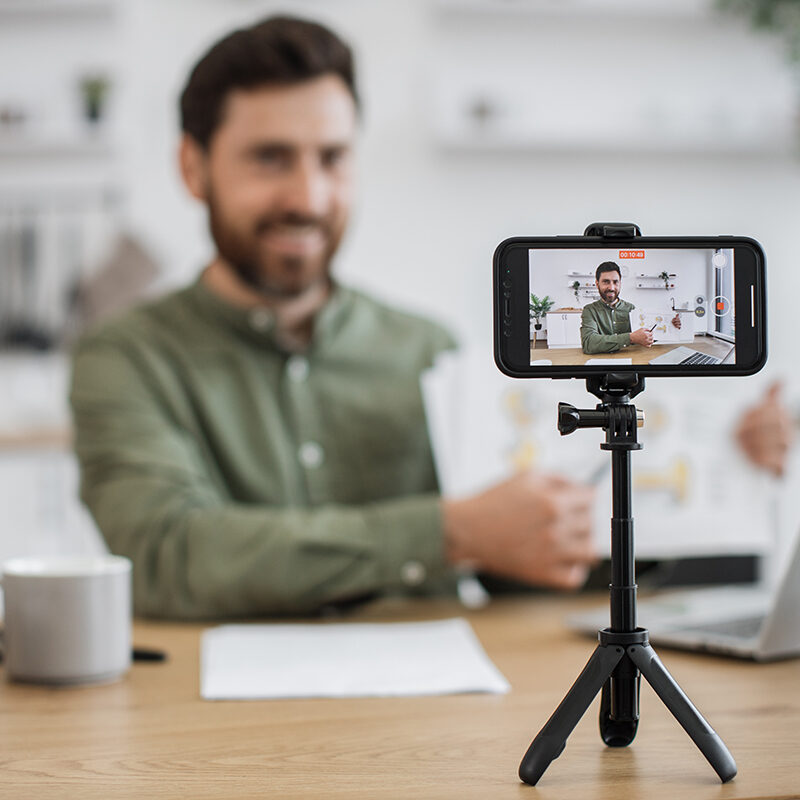 Blur background of caucasian man sitting at desk with laptop and showing financial report on smartphone camera. Competent financier making video lesson about reading and interpreting graphic charts.